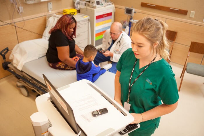 A nurse in green scrubs works on a computer cart at one of the renowned Minnesota Hospitals. Nearby, a doctor, part of the EPPA Health team, examines a young boy sitting on a hospital bed with his mother beside him.