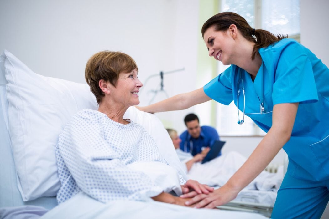 A nurse in blue scrubs smiles at a patient in a hospital bed while holding her hand, embodying the compassionate care for which Minnesota Hospitals are known. Two medical staff are engaged in conversation in the background.