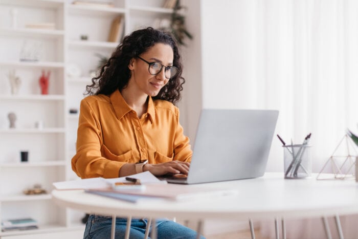 A woman with glasses and a yellow blouse is sitting at a white table, using her laptop in a bright room with shelves in the background. She appears focused, possibly researching about EPPA Health or Emergency Room Physicians, amidst the bustling environment reminiscent of Minnesota Hospitals.