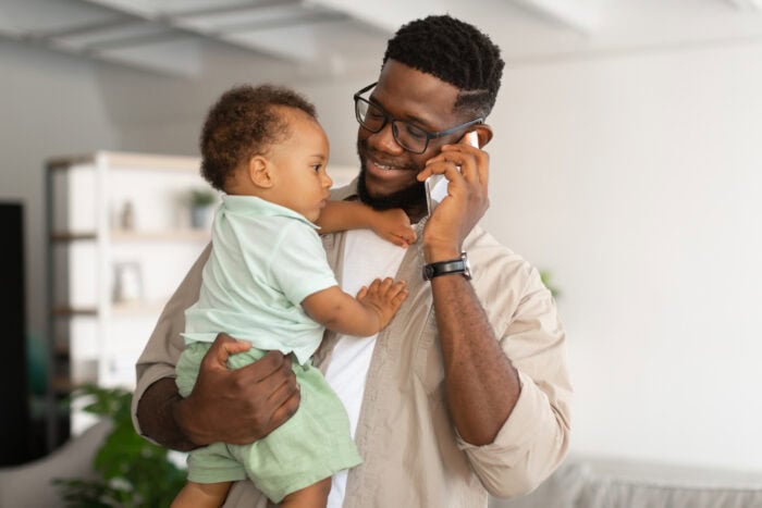 A man, likely an emergency room physician from EPPA Health, is holding a toddler while talking on a smartphone indoors. The man is smiling warmly as the toddler reaches out toward him, bringing a touch of joy to a moment that reflects the balance many Minnesota hospitals inspire.