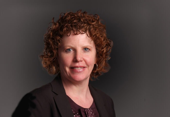 A person with curly hair and wearing a dark blazer smiles against a plain dark background, embodying the professionalism of Minnesota Hospitals.