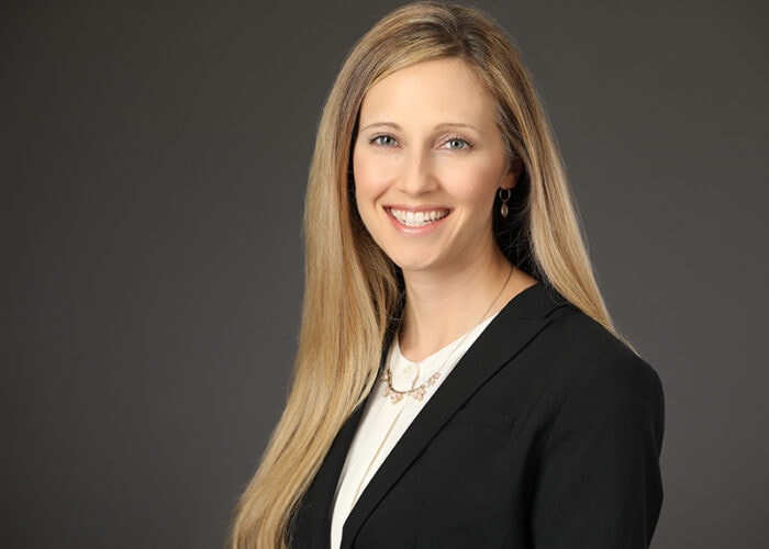 A person with long blonde hair, dressed in a black blazer and white shirt, smiles confidently against a gray background, reminiscent of the professionalism found in Minnesota hospitals like EPPA Health.