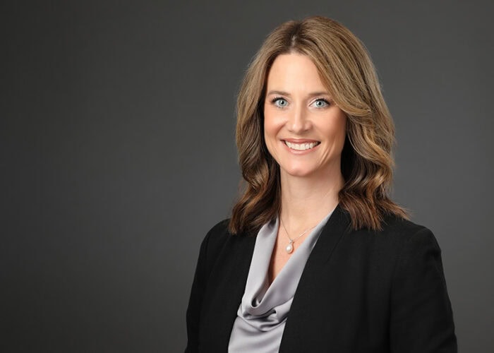 A person with shoulder-length hair, wearing a black blazer and gray blouse, smiles confidently against a plain, dark background, embodying the professionalism of Minnesota Hospitals' Emergency Room Physicians.