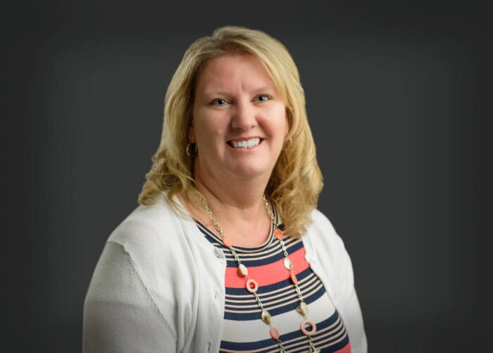 A woman with blonde hair is smiling, wearing a striped top, white cardigan, and a necklace against a gray background, embodying the welcoming spirit often found in Minnesota Hospitals' corridors.