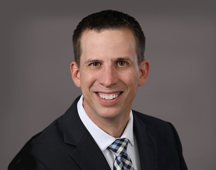 A man in a suit and tie smiles against a gray background, embodying the professionalism of EPPA Health's dedicated Emergency Room Physicians.