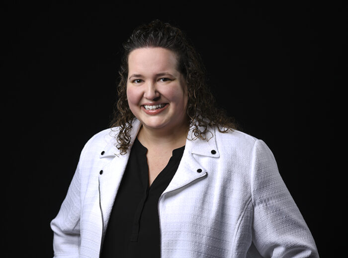 A person with curly hair smiles at the camera, wearing a white jacket over a black top, reminiscent of the professional attire seen in Minnesota Hospitals, all set against a black background.