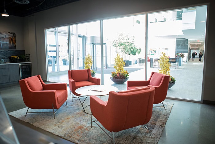 A modern office lounge with four red chairs around a small circular table on a rug, near large windows that overlook a bright corridor. The space reflects the innovative design ethos of EPPA Health, where emergency room physicians often gather for collaborative discussions.